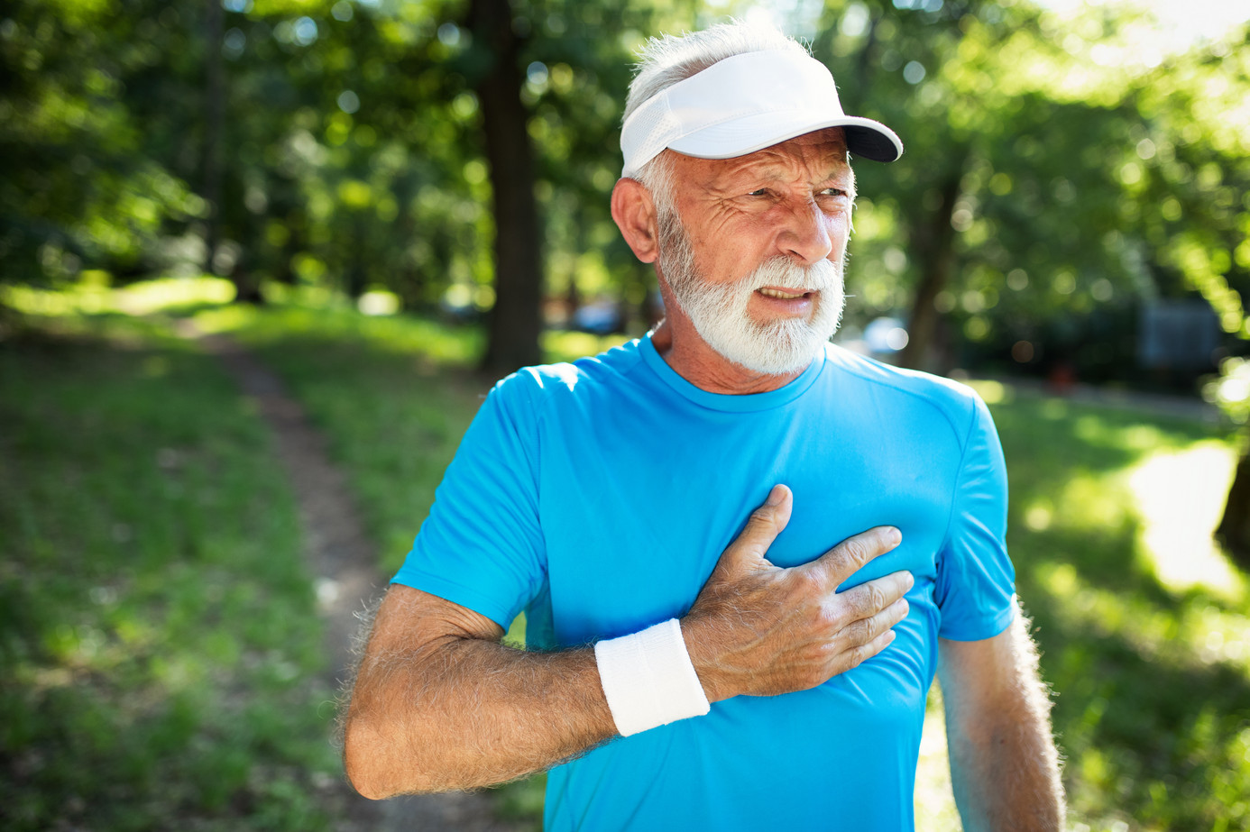 Älterer Mann in Sportkleidung in der Natur greift sich mit verzerrtem Gesicht ans Herz