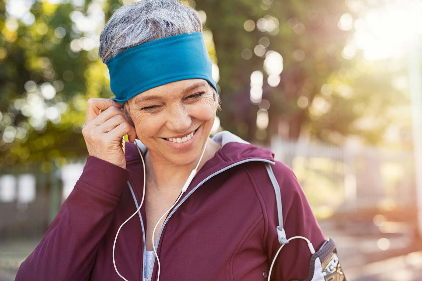 Frau mittleren Alters in Sportkleidung in der Natur steckt sich fröhlich Kopfhörer, die zu einem Handy führen, in die Ohren