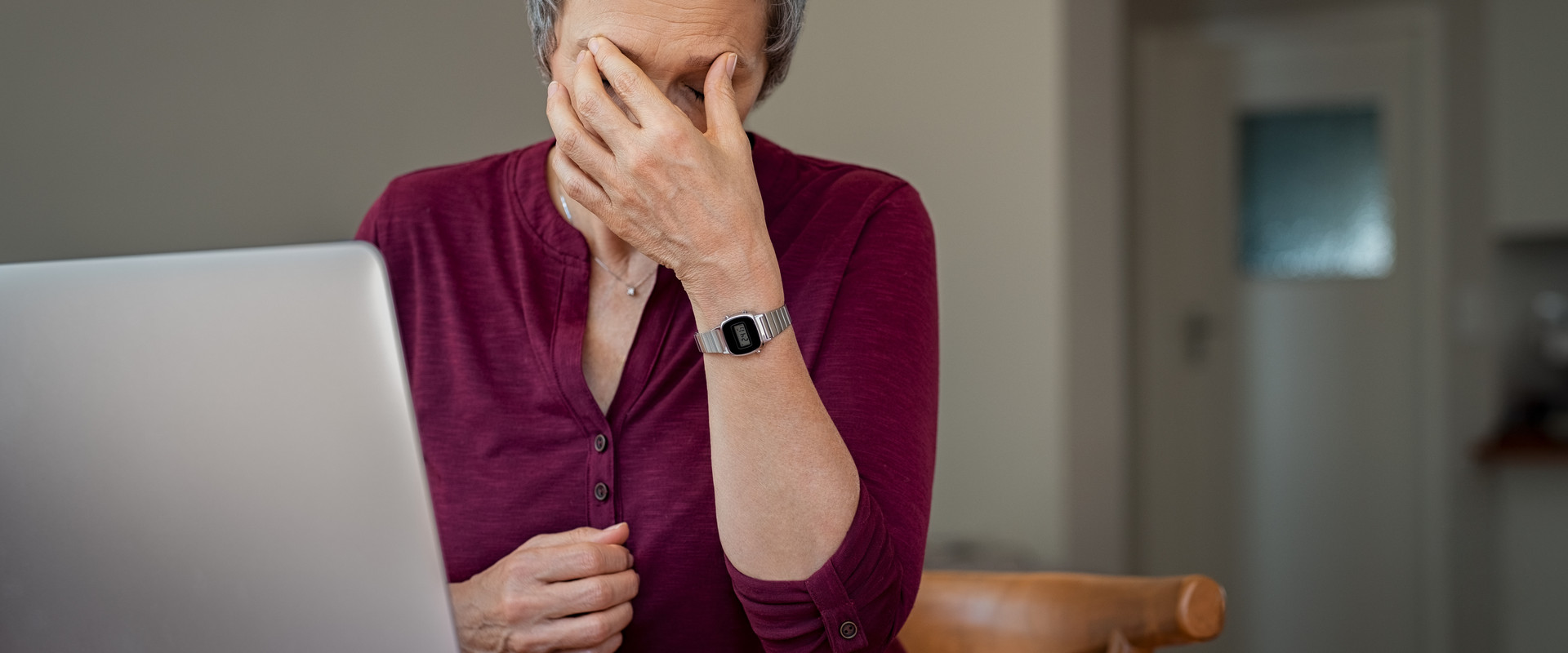 Frau mittleren Alters sitzt am Tisch, vor sich ein aufgeklapptes Laptop und hält sich eine Hand vor das Gesicht