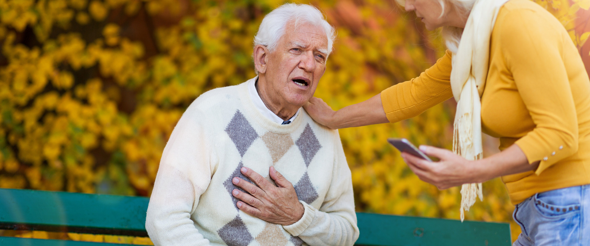 Älterer Mann sitzt im Park auf einer Bank, fasst sich ans Herz, eine Frau mit Handy in der Hand legt eine Hand auf seine Schulter